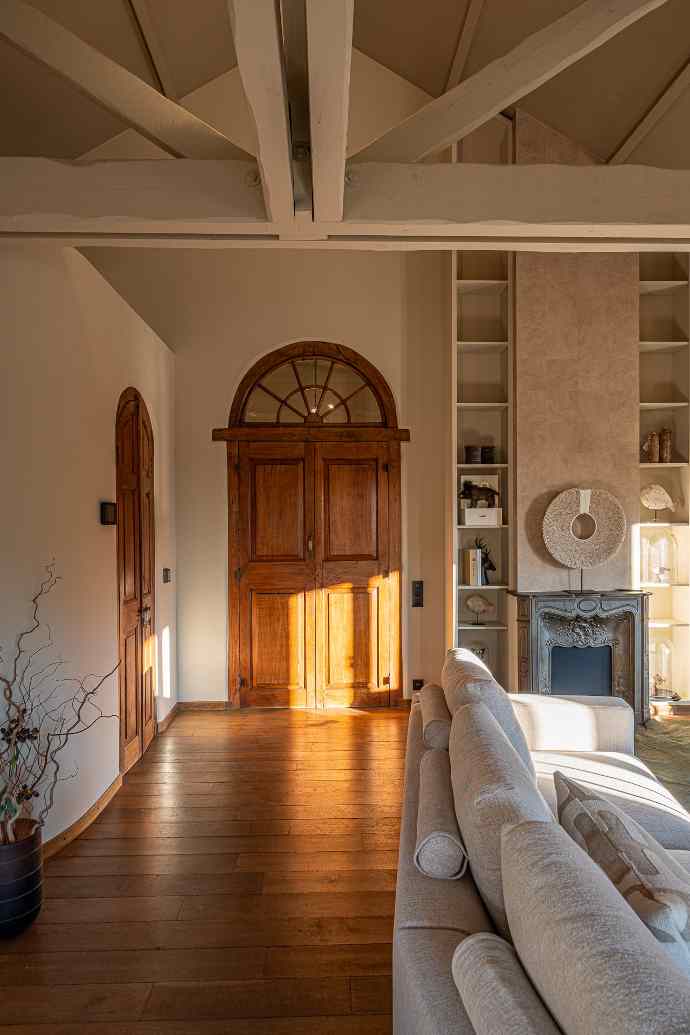 Salon chaleureux avec porte en bois, canapé, cheminée, et poutres blanches au plafond